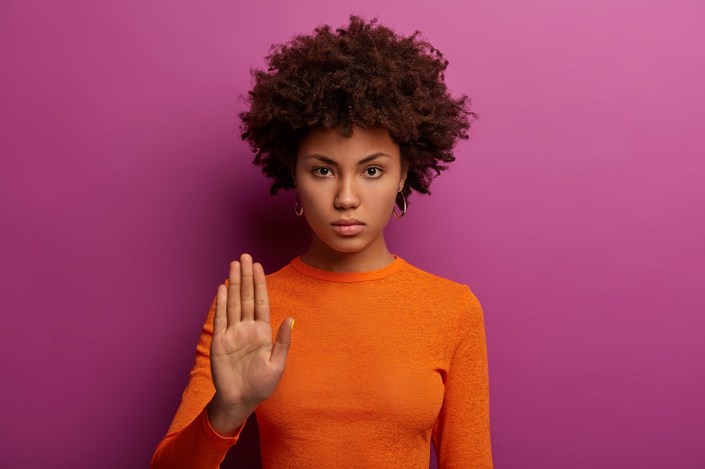 Foto de uma mulher negra encostada em uma parede rosa faz o gesto de pare com a mão
