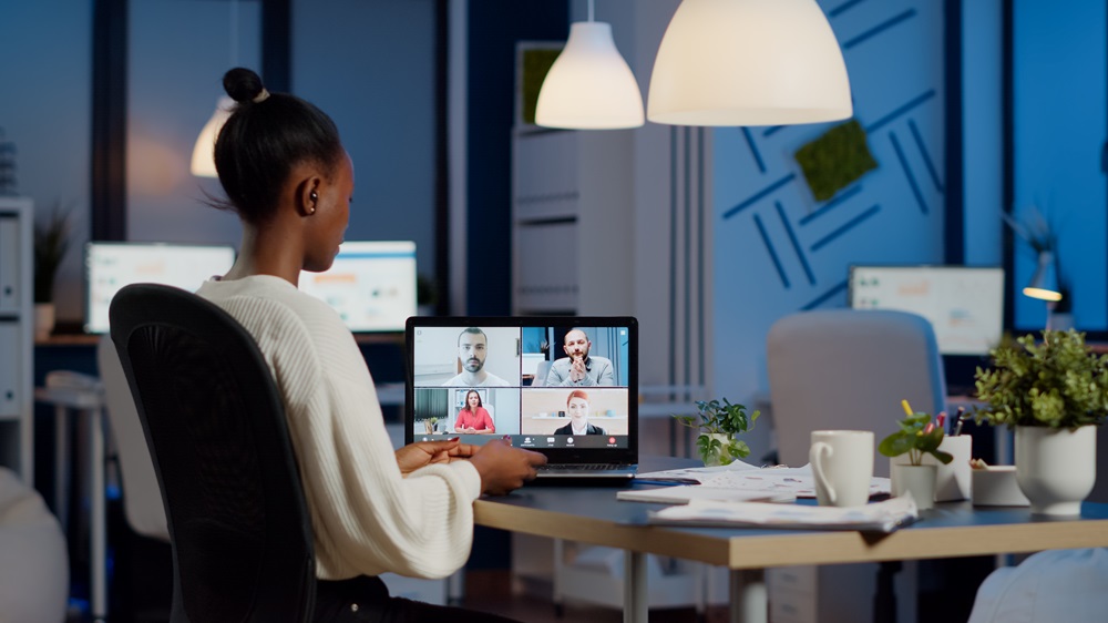 Foto de uma mulher negra sentada, em uma sala, e na sua frente há uma mesa e sobre ela um computador. Na tela, há uma chamada de vídeo em andamento com a participação de quatro pessoas.