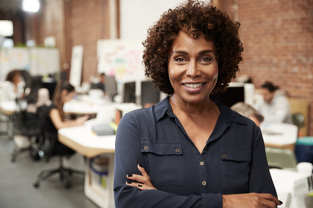 Foto posada de uma mulher negra em um escritório. Ela está com os braços cruzados e sorri.