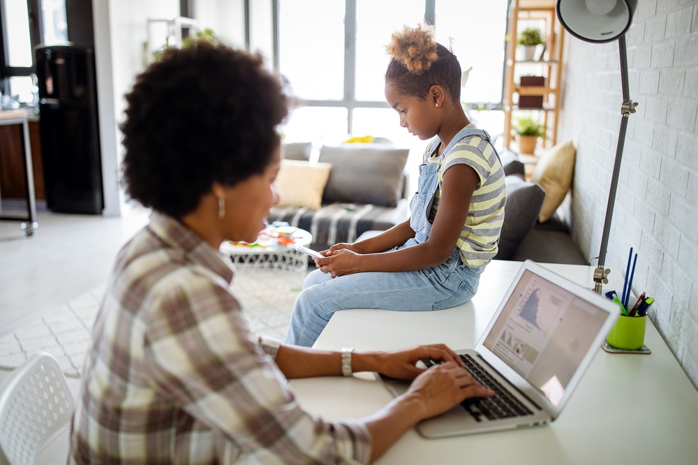 Foto de uma mulher negra trabalhando em um laptop enquanto sua filha está sentada ao seu lado e usa um celular.