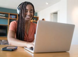 Foto de jovem negra com fones de ouvido sentada à mesa com as mãos no teclado do laptop que está sobre a mesa