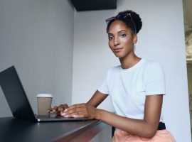 Foto de mulher negra jovem sentada em frente a um laptop com um copo de café ao lado do equipamento.