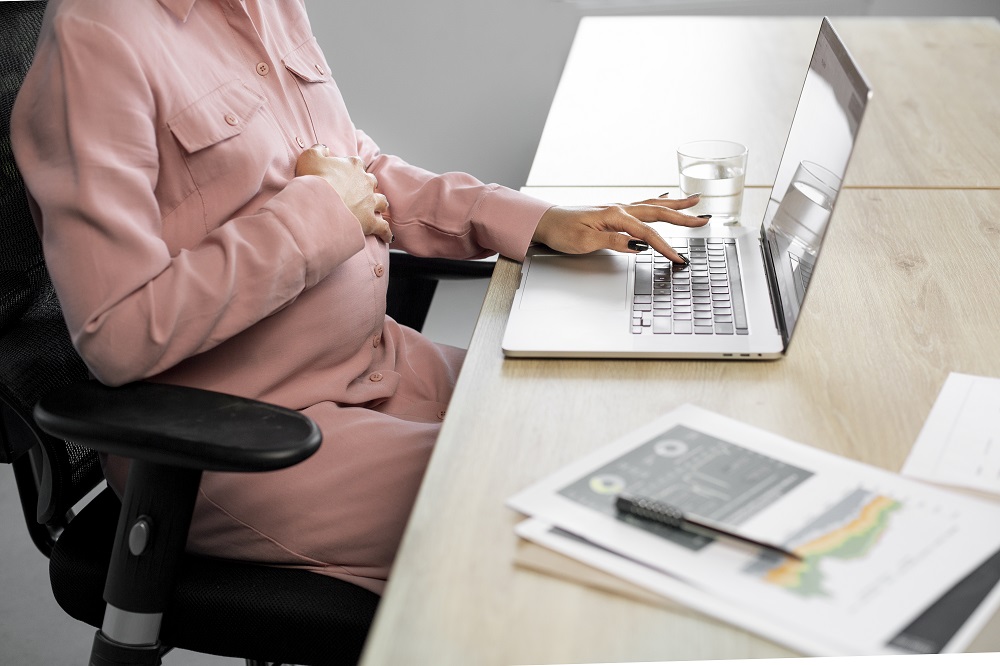 Foto em close de uma mulher grávida sentada com a mão direita apoiada em cima da barriga, enquanto a esquerda está em cima do teclado de um laptop.