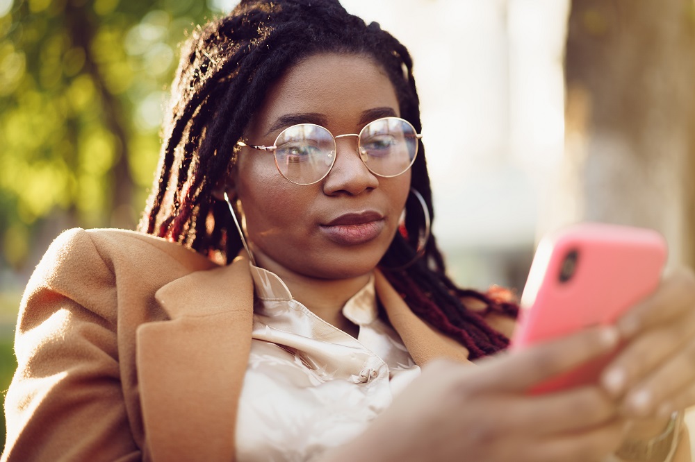 Foto de uma mulher negra sentada em uma área externa enquanto mexe com um celular.