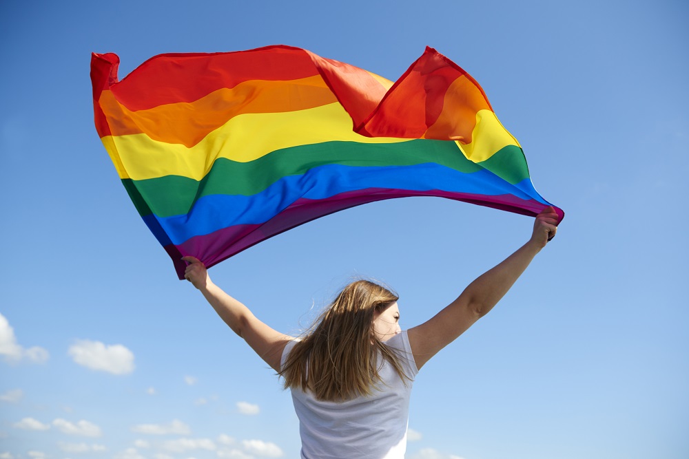 Foto de uma mulher de costas com os braços levantados e segurando a bandeira do Orgulho LGBTQIA+.