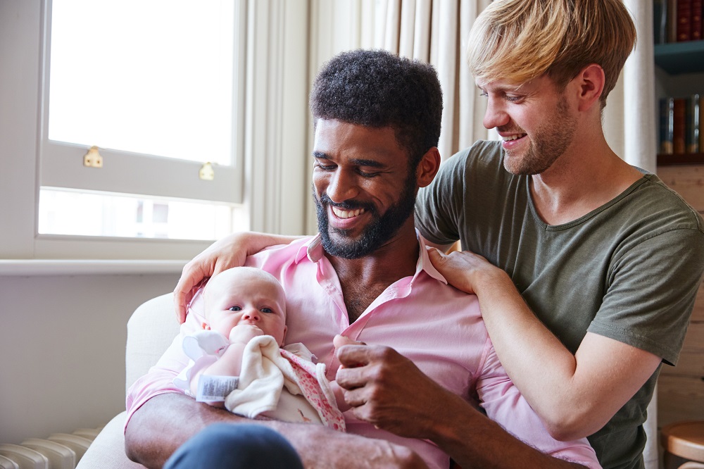 Foto de um casal de pais gays sorrindo para a filha bebê que está no colo de um deles, que segura a mãozinha esquerda da criança enquanto ela coloca a mão direita na boca.