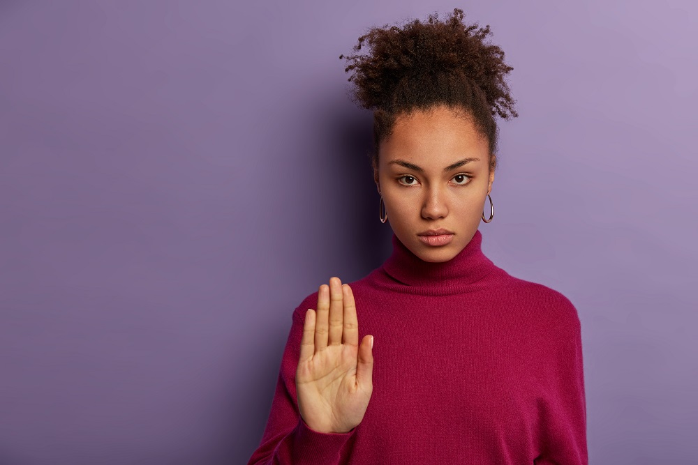 Foto posada de uma mulher negra jovem com uma expressão séria. Ela está com o braço esquerdo levantado mostrando a palma da mão para a câmera.