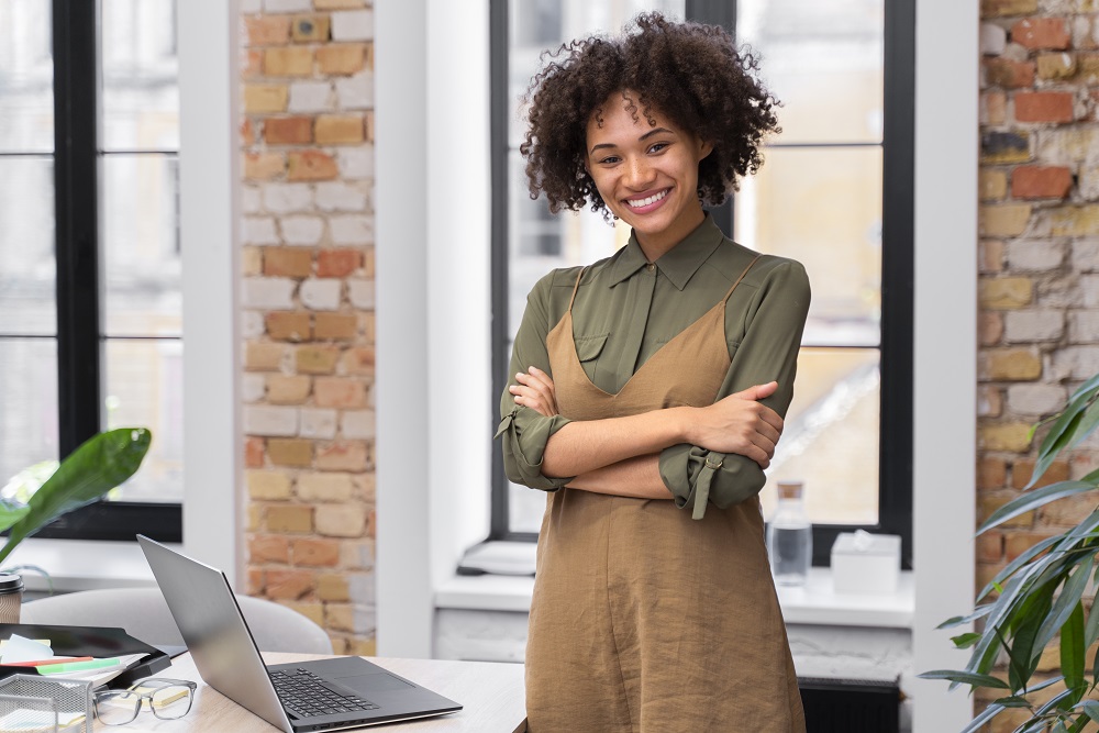 Foto de uma mulher negra, em pé, em um escritório com os braços cruzados. Ela sorri