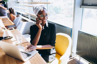 Foto de uma mulher negra sentada em uma cafeteria. Ela fala ao telefone enquanto a mão esquerda está em cima do mousepad do laptop