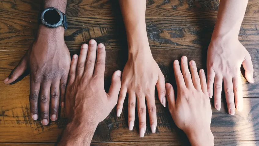 Foto de quatro mãos, de diferentes tons de pele, uma ao lado da outra sobre uma superfície de madeira