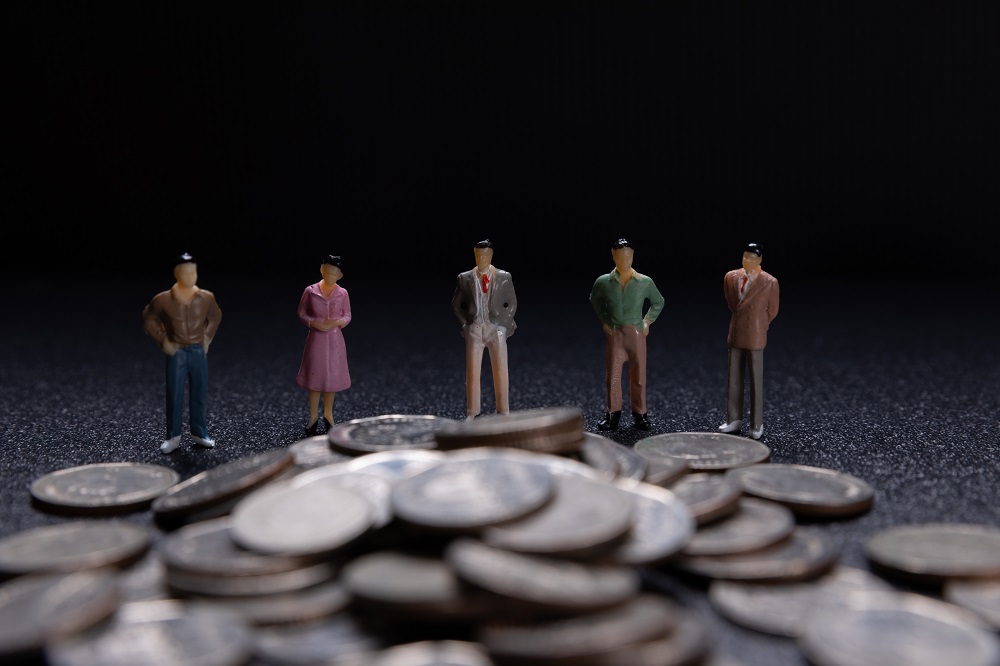 Foto de quatro bonecos em miniatura de homens e uma mulher, todos colocados em pé, lado a lado, e na frente deles há uma pilha de moedas