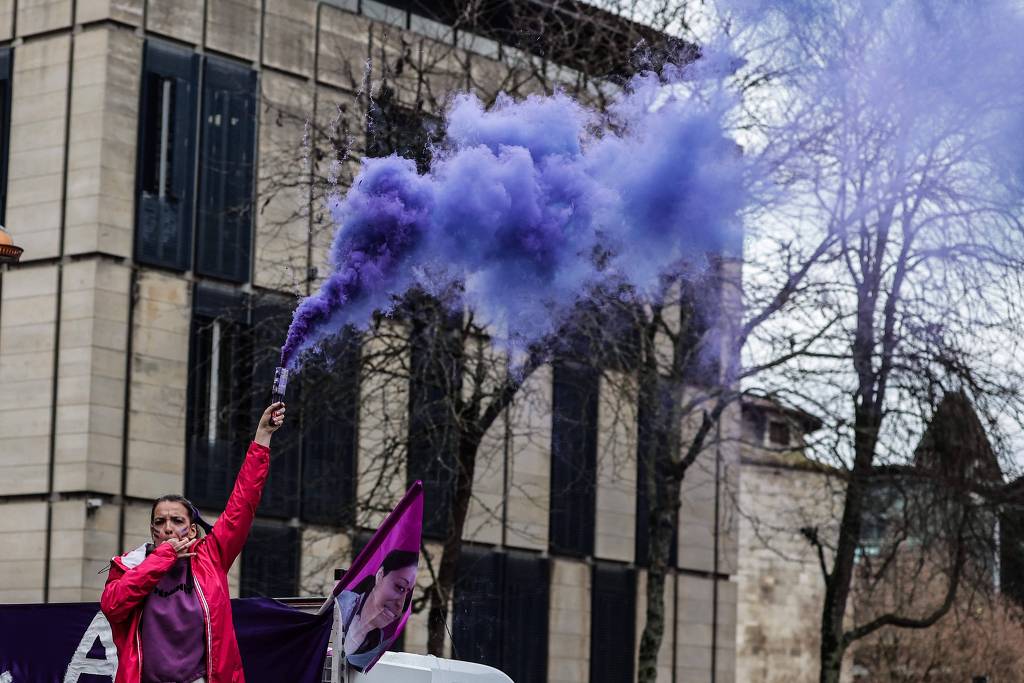 Foto de uma mulher em Bordeaux, na França, durante um protesto. Ela está com o rosto pintado com linhas paralelas lilás, está assoviando com a mão direita enquanto a esquerda segura um objeto que solta fumaça lilás