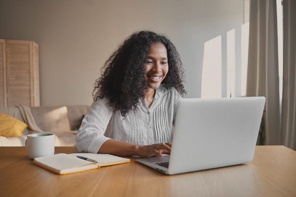 Foto de uma mulher negra jovem em uma sala, sentada, com as mãos sobre o teclado de um laptop.