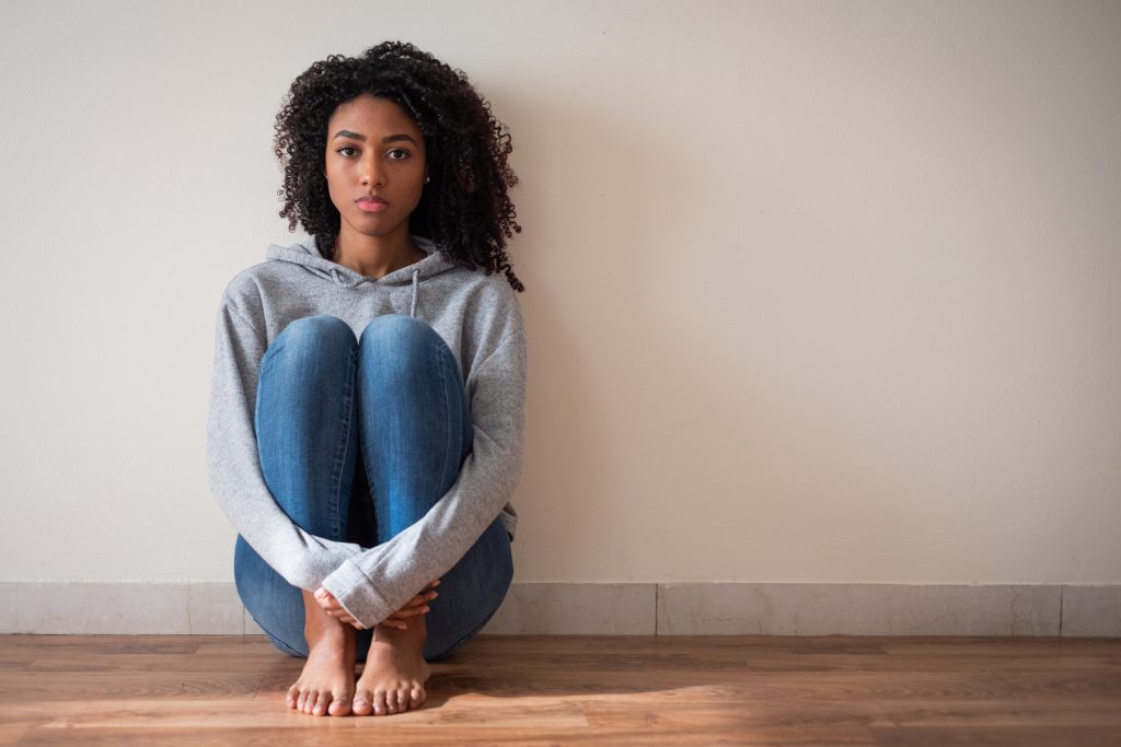 Foto de uma mulher negra jovem sentada no chão e encostada em uma parede, com as pernas próximas ao peito e as mãos abraçando as pernas