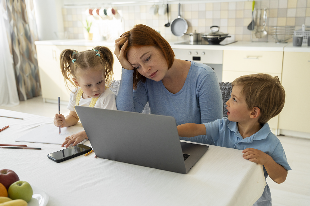Foto de uma mãe trabalhando em um laptop na cozinha, enquanto está com a filha e o filho ao redor. Ela está com uma das mãos na cabeça e uma expressão de cansada