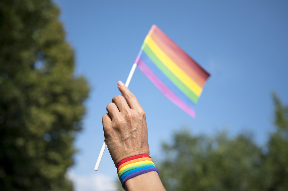Foto da mão de uma pessoa segurando uma bandeira com as cores do orgulho LGBTQIA+. No pulso, há uma pulseira com as cores do arco-íris. Ao fundo, há um céu azul e algumas árvores.