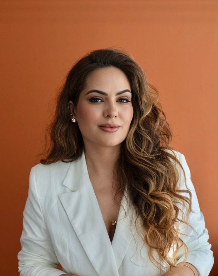 Foto de Mariana Covre em frente a uma parede. Ela é branca tem cabelos longos ondulados, veste um blazer branco e sorri levemente.