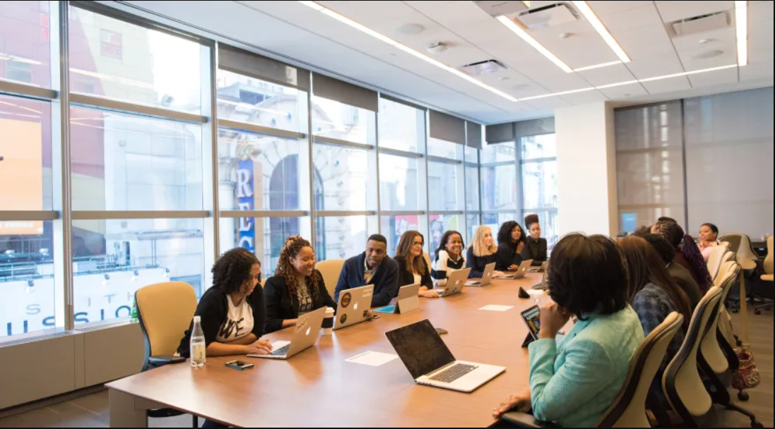 Foto de um grupo diverso de pessoas sentadas ao redor de uma mesa em uma sala de reuniões. Há laptops e tablets sobre a mesa.