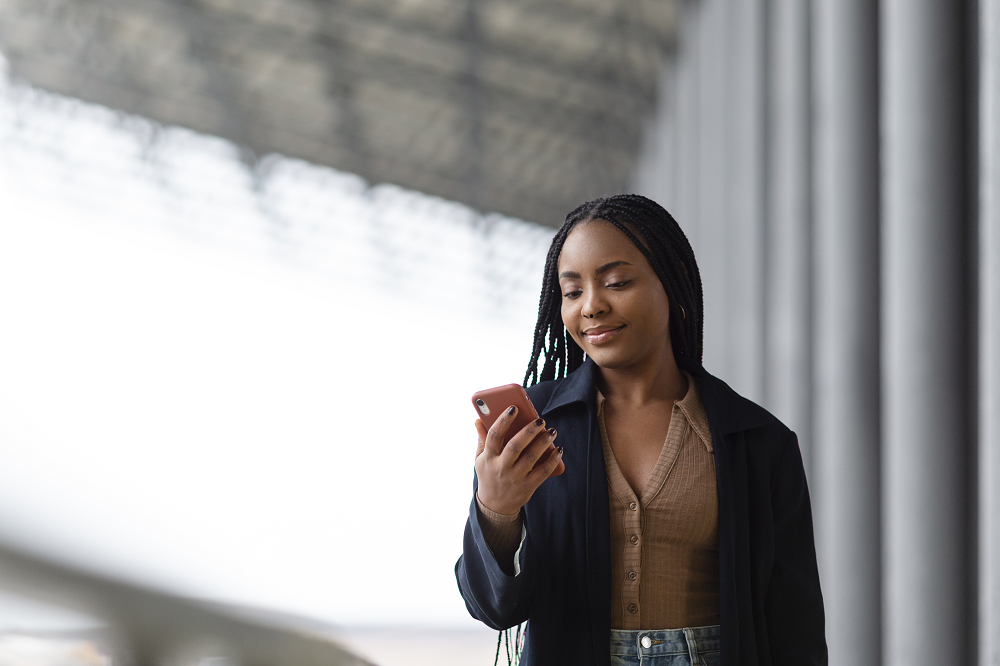 Foto de uma jovem negra olhando para o celular que está em sua mão esquerda. Ela sorri.