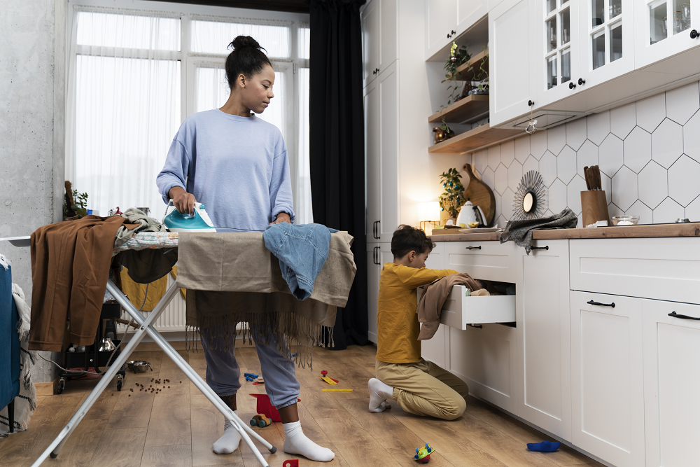 Foto de uma mulher e uma criança no interior de uma cozinha. Ela, em pé, passa a roupa, enquanto o menino mexe em uma gaveta.