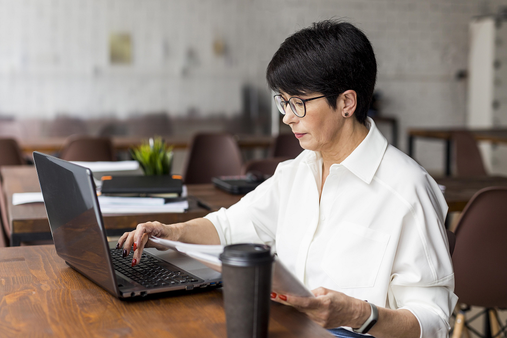 Foto de uma mulher oriental de perfil. Ela está sentada em uma sala e está com uma das mãos sobre o teclado de um laptop.