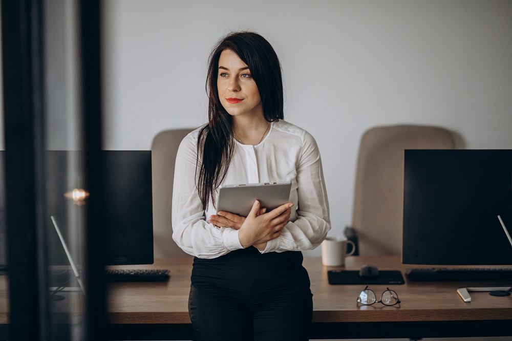 Foto de uma mulher em um escritório. Ela está em pé e segura um tablet nas mãos