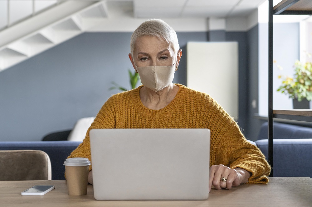 Foto de uma mulher idosa, de cabelos curtos, usando máscara, enquanto usa um laptop