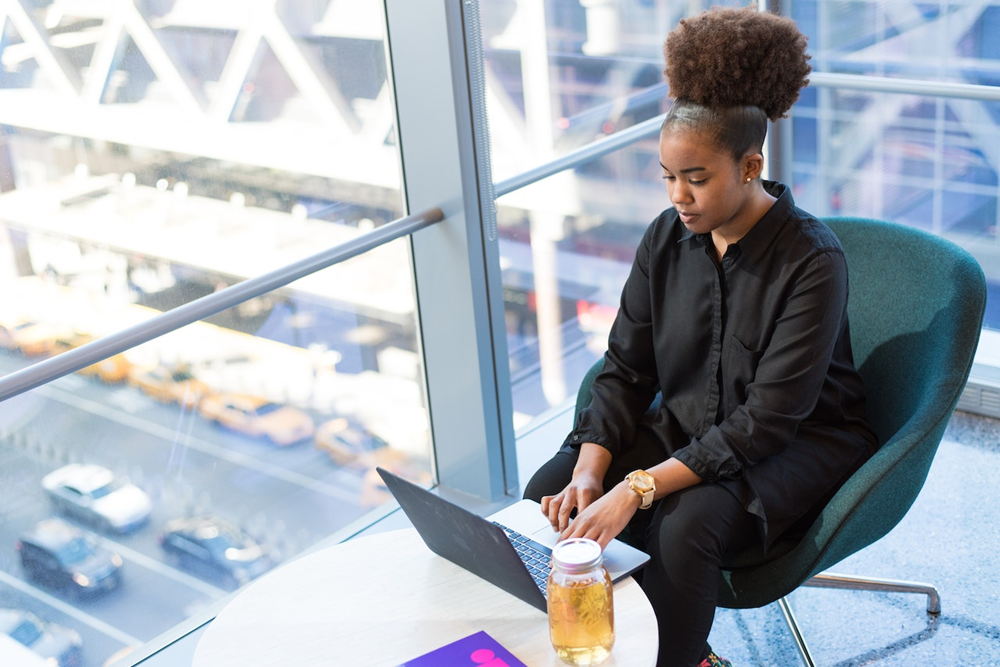 Foto de uma mulher negra sentada em uma sala com paredes de vidro. Ela digita no teclado de um laptop.