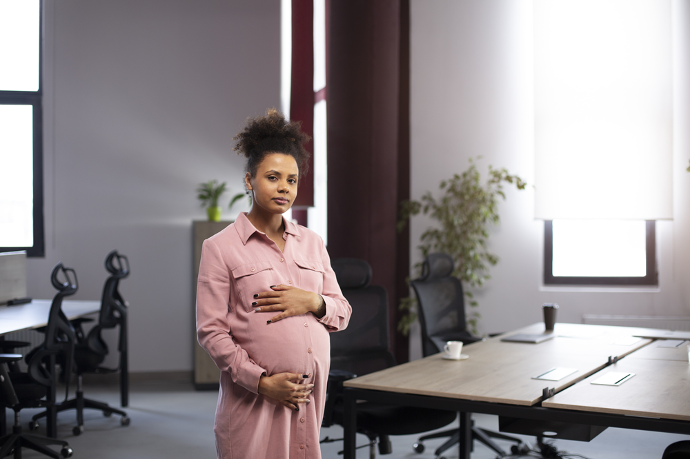 Foto de uma mulher negra grávida, em pé, com as mãos em cima da barriga.
