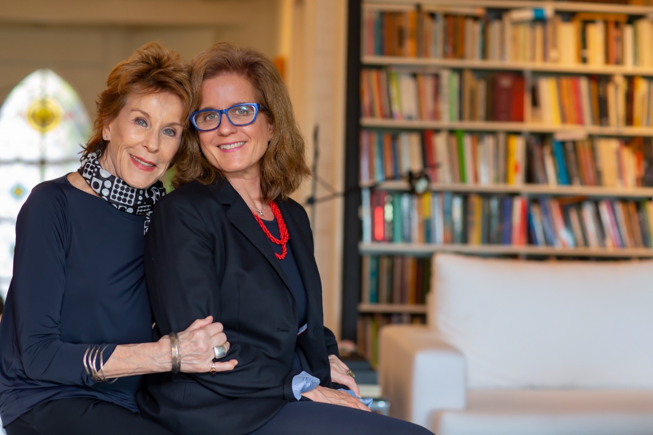Foto posada de Sylvia Loeb e Carla Leirner sentadas em uma sala com uma estante cheia de livros.