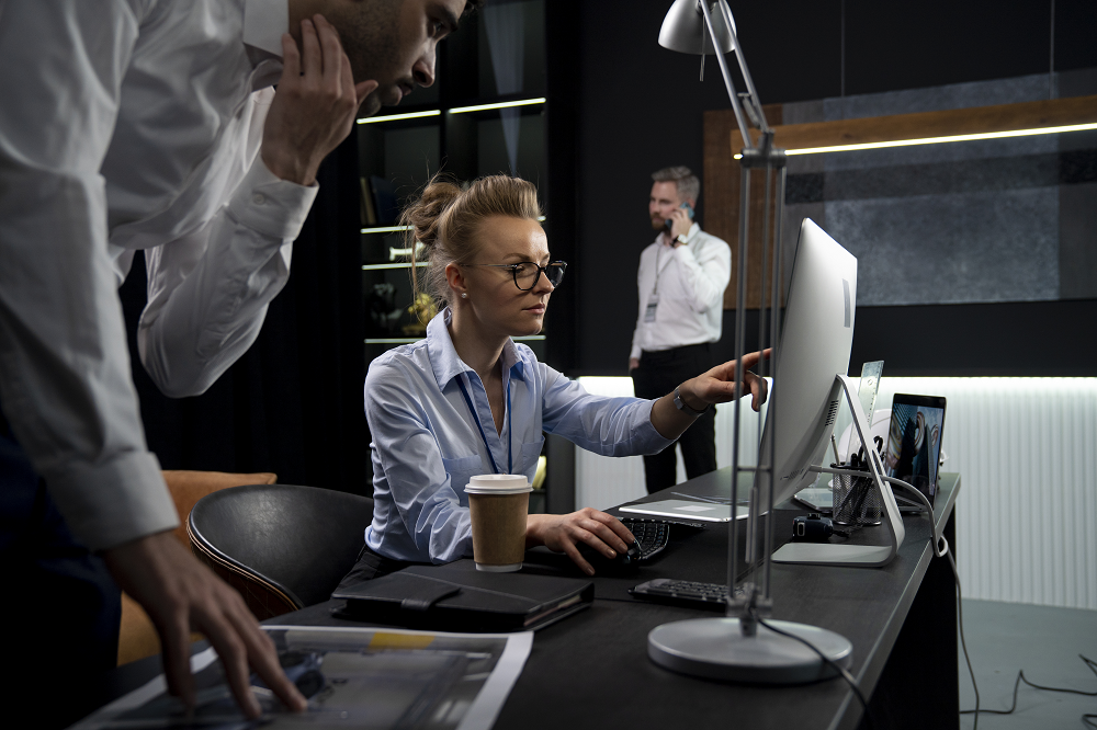 Foto de uma mulher sentada em um escritório. Ela aponta o dedo indicador direito para a tela de um computador de mesa, e ao seu lado há um homem.