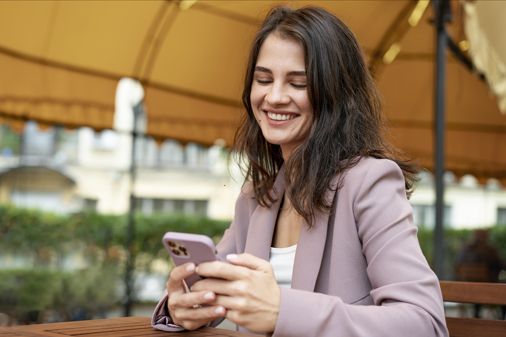Foto de uma mulher em uma área externa. Ela está sentada, mexendo no celular e sorri enquanto olha para o aparelho.