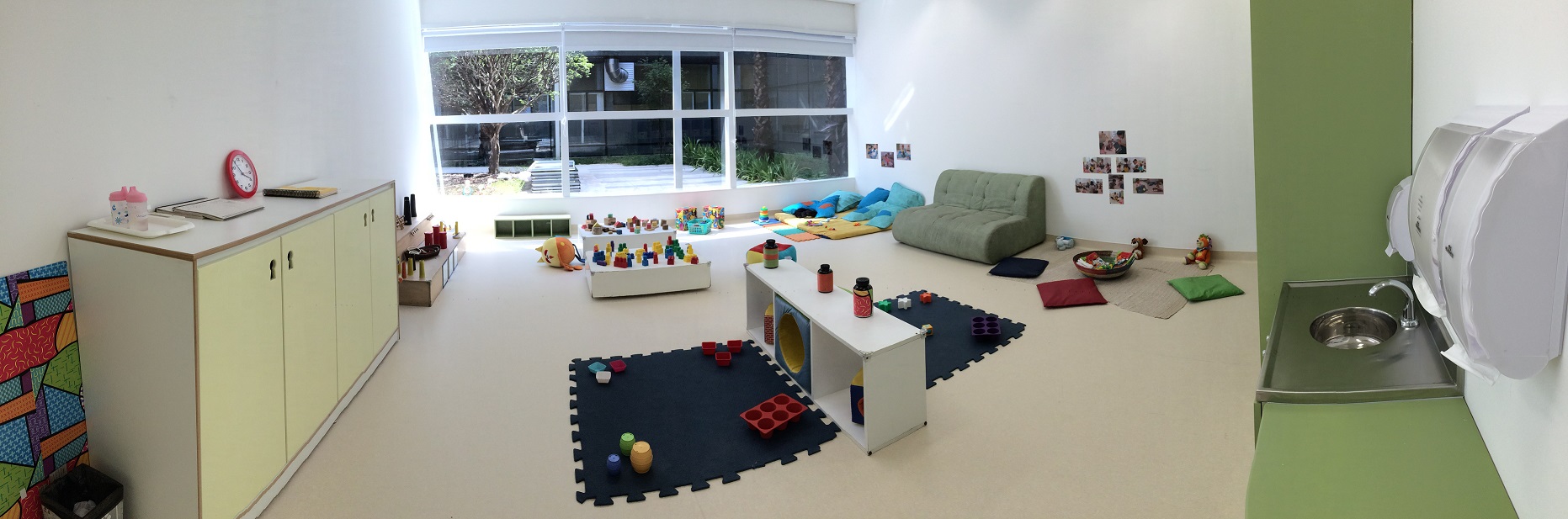 Foto em estilo panorâmico de uma sala reservada para ser o berçário. Há um sofá e vários brinquedos no chão.