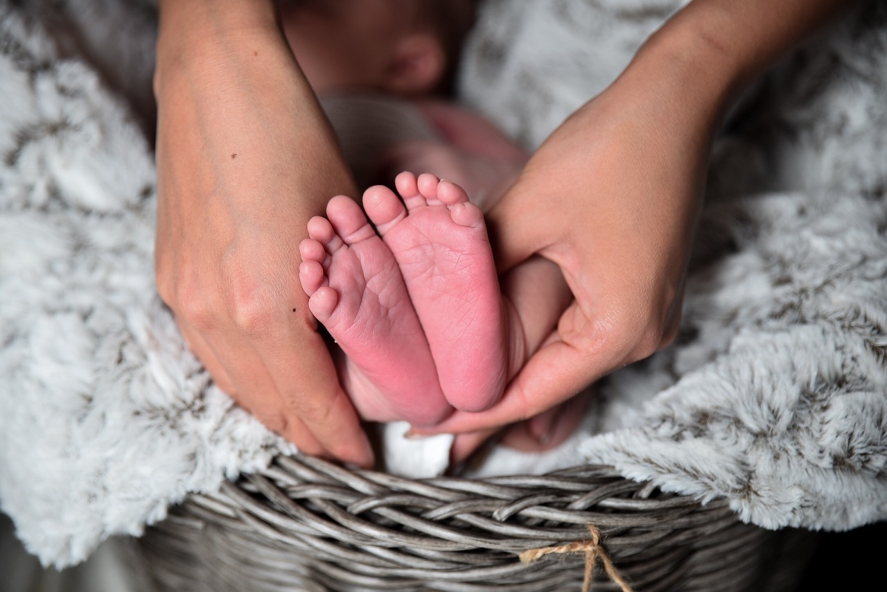 Foto em close dos pés de uma criança recém nascida. Ao seu redor, estão as mãos de uma pessoa adulta. A criança está em uma cesta com uma manta.