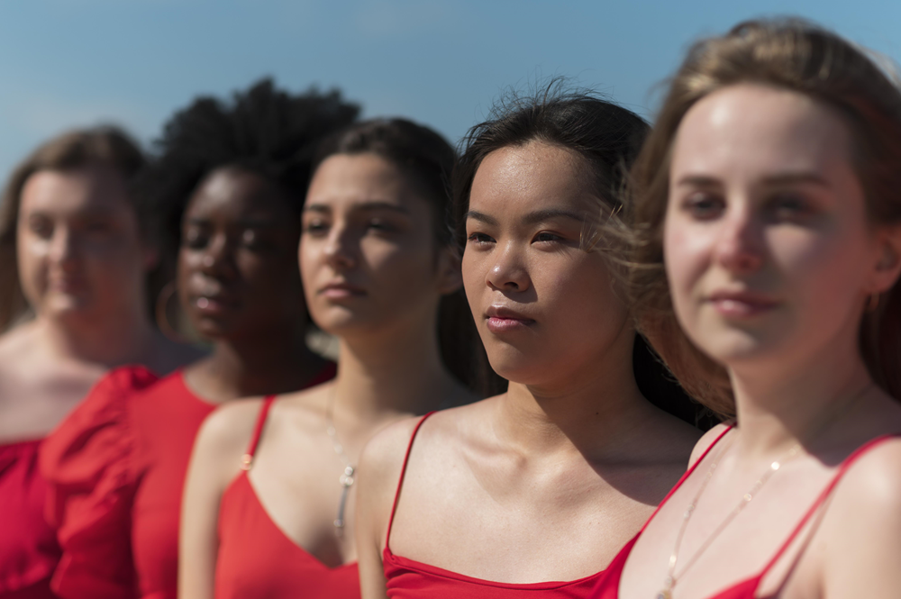 Foto de cinco mulheres, em pé, lado a lado, em um ambiente aberto. Todas vestem roupas vermelhas e estão com uma expressão séria no rosto.