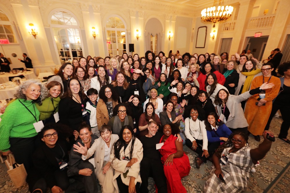 Foto de um grupo diverso de mulheres, sendo que algumas estão sentadas no chão e outras em pé, em um salão do The Yale Club of New York City.