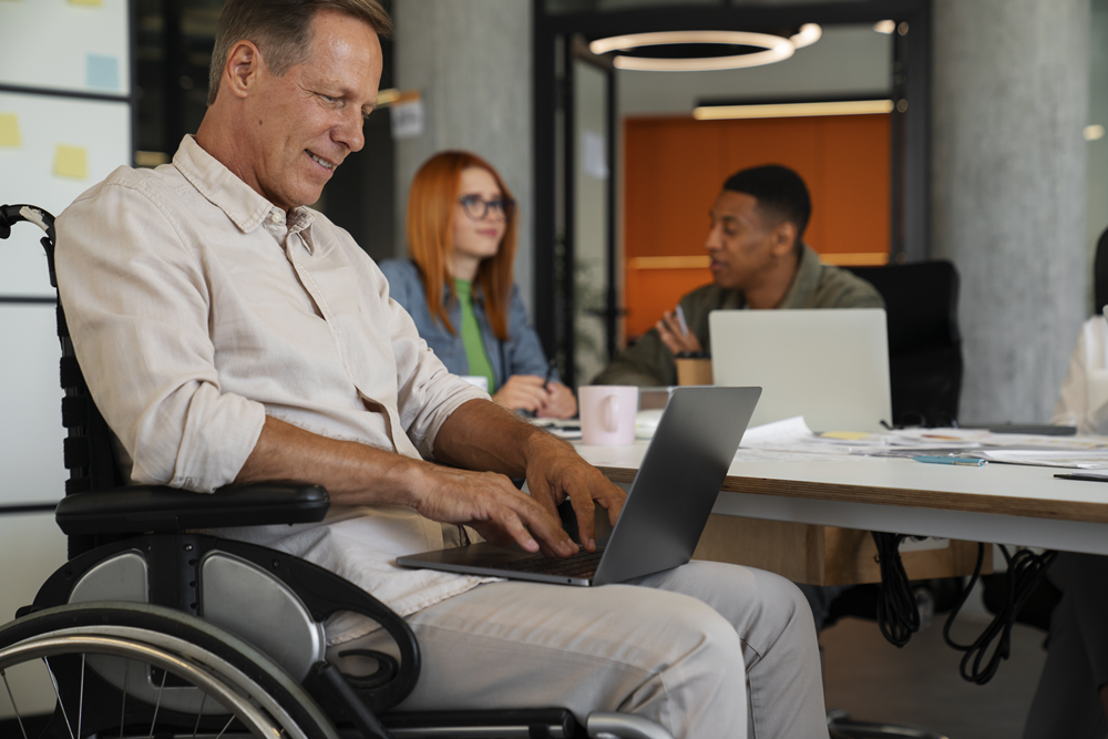 Foto de um homem cadeirante com um laptop no colo, e ao fundo há uma mulher e um homem conversando.