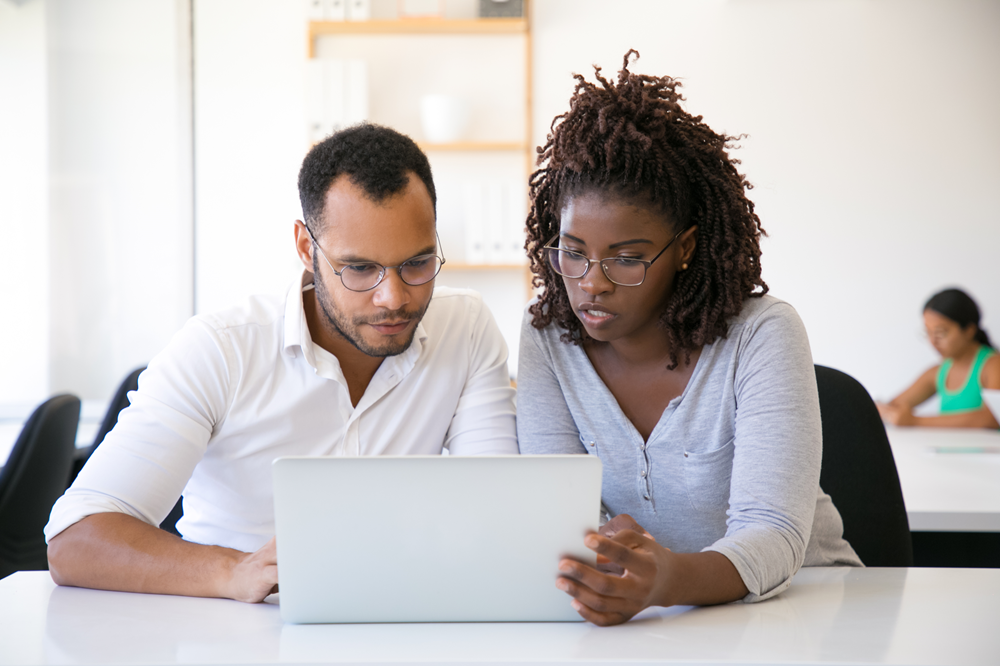 Foto de um homem e uma mulher em um escritório. Estão sentados um do lado do outro e olham para a tela de um laptop.