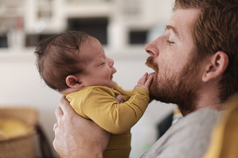 Foto de perfil de homem segurando um bebê na altura do seu rosto. A criança coloca as mãos em na boca do homem.