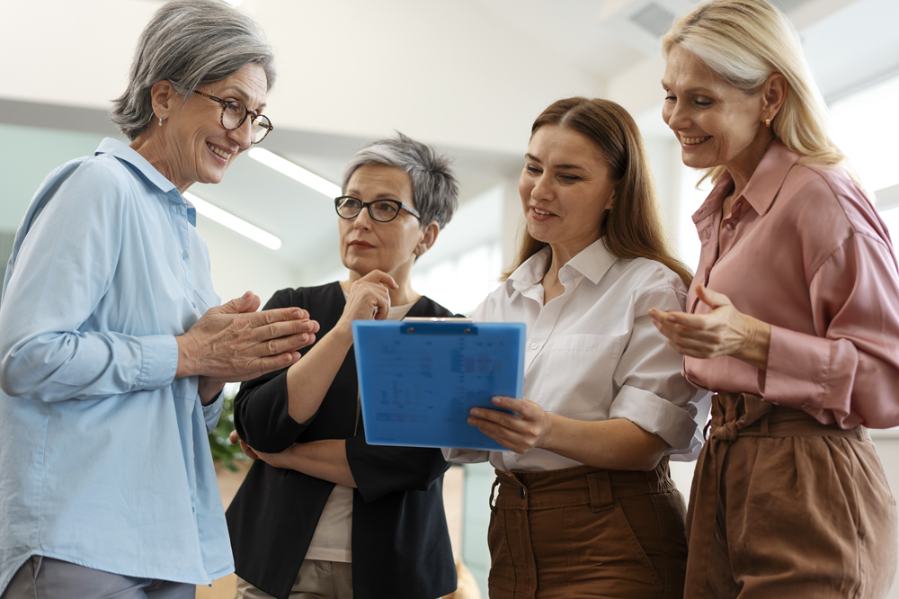 Foto de um grupo de quatro mulheres de diferentes idades conversando em uma roda. Uma delas, segura uma prancheta em uma das mãos.
