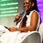 Foto de Mafoane Odara, mulher negra com cabelos longos trançados. Fala ao microfone enquanto faz a mediação do painel Cultura e Mídia.