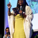 Foto de Benilda Brito, uma mulher negra com cabelos longos com tranças. Ela está com um microfone em cima do palco.