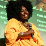 Foto de Carla Akotirene, mulher negra com cabelo black power. Fala ao microfone enquanto participa do painel Cultura e Mídia.