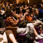 Foto de um grupo diverso sentado nas poltronas da plateia do teatro.