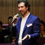 Foto de um homem, em pé, em meio a plateia, falando ao microfone.