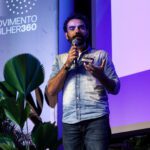 Foto de Guilherme Nascimento, homem branco com barba e bigode. Fala ao microfone no palco do evento.
