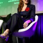 Foto de Paola Carosella, mulher branca com cabelos levemente ondulados. Ela está sentada em uma poltrona branca, que está colocada no palco para sua participação do painel Cultura e Mídia.