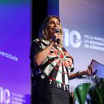 Foto de Maite Schneider, mulher trans branca com cabelos loiros na altura dos ombros. Ela fala ao microfone no palco do evento.