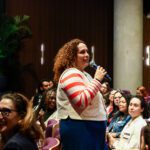 Foto de uma mulher, em pé, em meio a plateia, falando ao microfone.