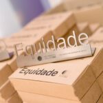 Foto de várias pilhas de caixas de papelão e sobre uma delas, um peso de metal com a palavra equidade escrita.